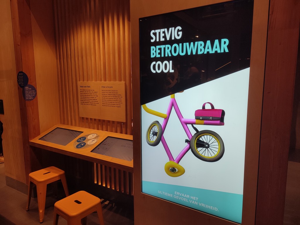 In the foreground and the right side of the image is what looks like a bicycle advertisement, with the Dutch words "Stevig, Betrouwbaar, Cool" written across the top. Below it is an AI generated image of a pink and yellow bicycle, though it looks very wrong. The main two wheels cannot touch the ground due to the sudden appearance of a third wheel below them. Below the bicycle is more Dutch, "Ervaar het ultieme gevoel van vrijheid." In the background, there are some tablets installed in a wooden panel with stools for people to sit at.