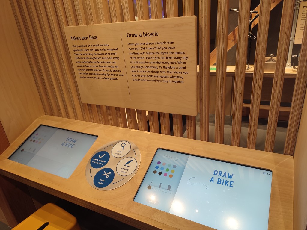 Tablets installed in wooden panels with stools in front of them. On the tablets reads the phrase, "Draw a bike." Above the tablets is a sign that reads in both English and Dutch, "Draw a bicycle. Have you ever drawn a bicycle from memory? Did it work? Did you leave anything out? Maybe the lights, the spokes, or the brake? Even if you see bikes every day, it's still hard to remember every part. When you design something, it's therefore a good idea to draw the design first. That shows you exactly what parts are needed, what they should look like and how they fit together."