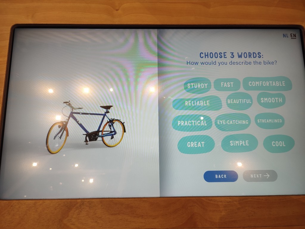 An AI image of a blue and yellow bicycle is on the left of the image. On the right, a prompt appears to "Choose 3 words: How would you describe the bike?" The words to be selected from are, "Sturdy, Fast, Comfortable, Reliable, Beautiful, Smooth, Practical, Eye-catching, Streamlined, Great, Simple, Cool." Below are options to either go back or go next.