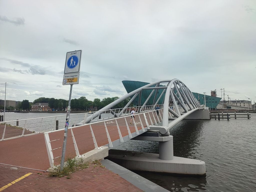 A pedestrian and cycle bridge over water, with a steel arching design over top. In the background is a large building that has a similar ship to a small boat, rounded and raised to the left.