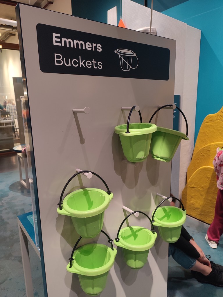 A small display stand with six green buckets hanging and nine total pegs to hang them on. The top of the display stand says, "Buckets" in both English and Dutch. The floor in the background is wet.