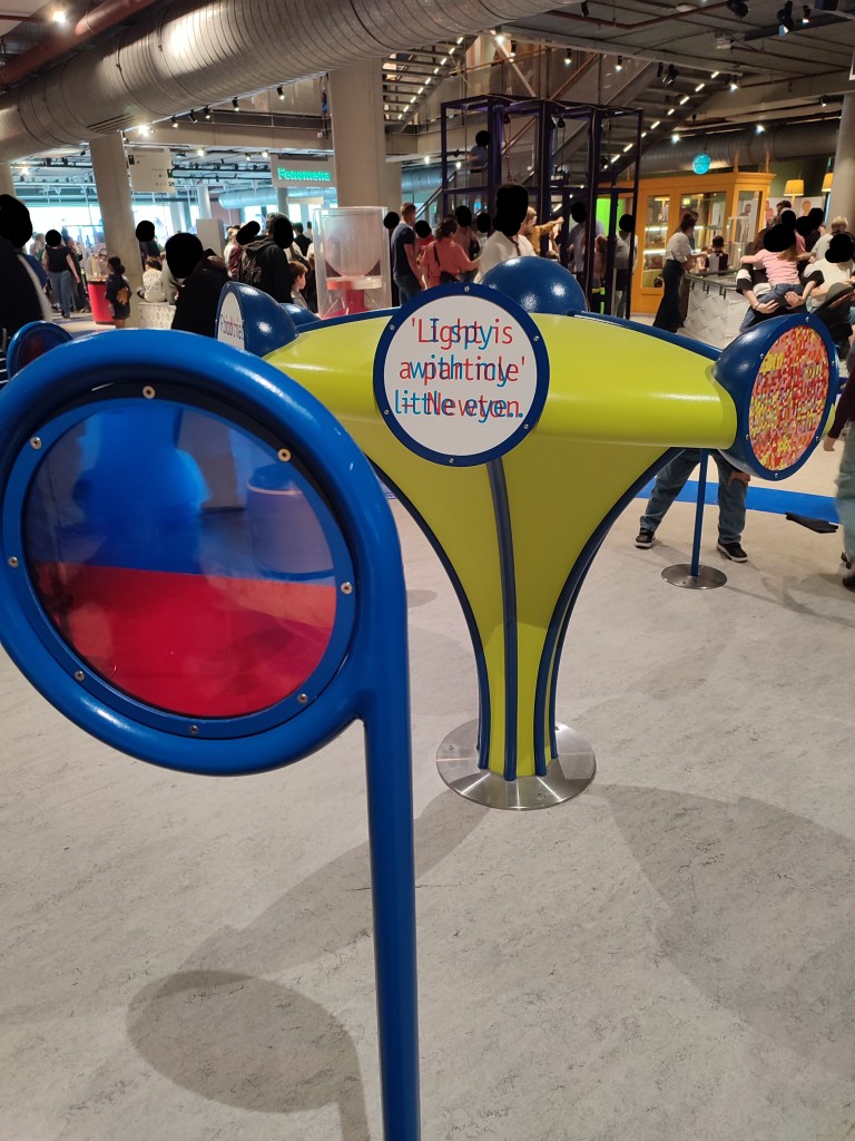 A yellow fixture sits in the middle of the frame. On it are several colour-based exhibits. The one most prominent features blue and red text overlayed on top of each other, making them almost illegible. Slightly in front of the fixture is a disc, half covered with blue and half covered with red, so when someone looks through the blue side they can only see the red text and vice versa. The red text reads, "'Light is a particle' - Newton" while the blue text reads "I spy with my little eye..".
