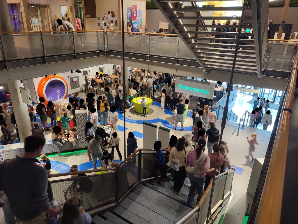 From a higher floor looking down a set of stairs, a large crowd has gathered near the base. Many people appear to be in a line, going from the left of the image and disappearing to a point that can't be seen behind part of the building structure. The entrance of the building is visible nearby.