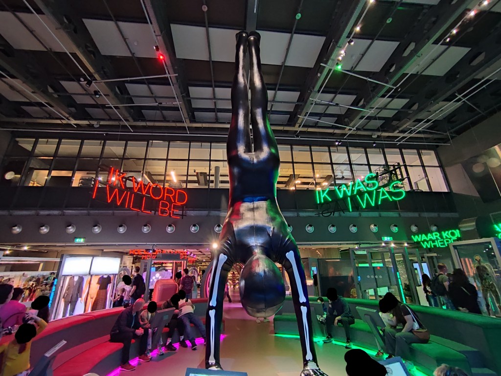 A large model of a human doing a handstand reaches from the floor to the ceiling, with arm and hand bones visible on top of a full body black covering. Around it are a few couches with screens in front of them, with some people looking at them, some with headphones. To the right of the model, a large green neon sign says, in English and in Dutch, "I was". To the left of the model, a large red neon sign says, in English and in Dutch, "I will be".