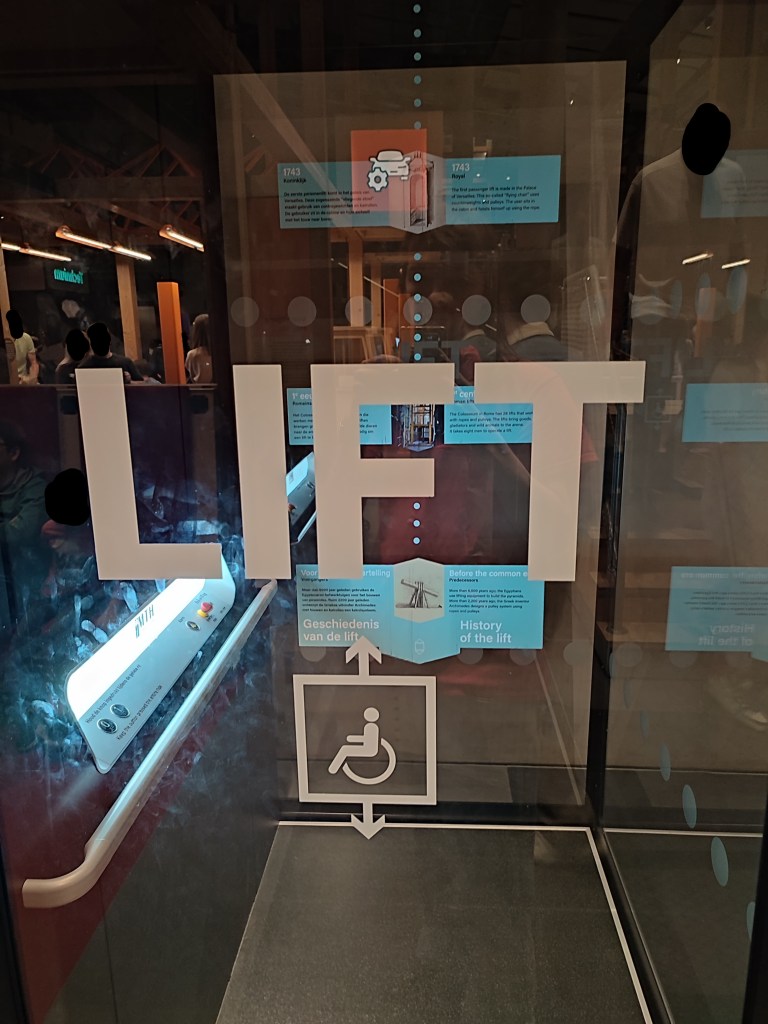 A glass elevator sits in front of the image, with the word "LIFT" in large block capitals and a diagram of a wheelchair user, indicating the purpose. On the back of the elevator is text and an exhibit that reads "History of the lift" with dots leading up to various pieces of text that progress through the timeline, though the text is largely illegible.