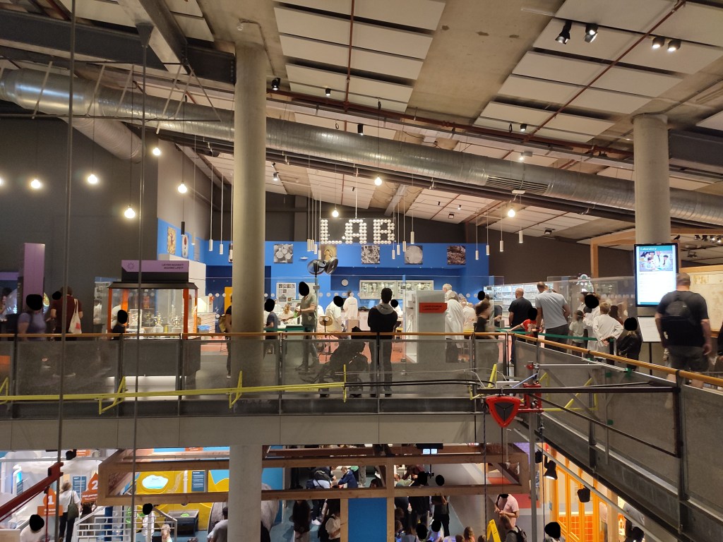 Across a small gap in the floor with railings, a section of venue is walled off with glass and labelled as "Lab". Many people can be seen inside of the lab, all wearing white lab coats. The entrance to the right of the image has a small line that has formed of people waiting to get in. A number of other people are watching what is happening inside the Lab. A large crowd can be seen on the floor below.