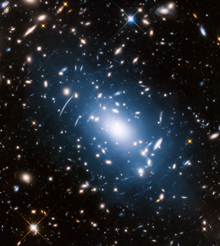 A black field showcasing an image filled with galaxies and stars. In the centre, a globular cluster fills the screen like a white smudge. Around it, galaxies appear to be warped and stretched unnaturally as the light is curved by gravity.