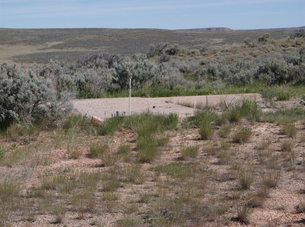 A dry landscape with the barren foundation of a building in the centre. Dry brush is scattered from the foreground to the horizon in a treeless landscape.
